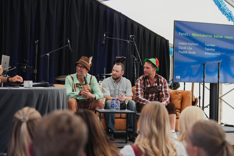 Två presentatörer på scenen talar i mikrofon under en 10-årsjubileumsfest i Sverige, med en stor presentationsskärm, evenemangssittning och blått och vitt ljus i bakgrunden.